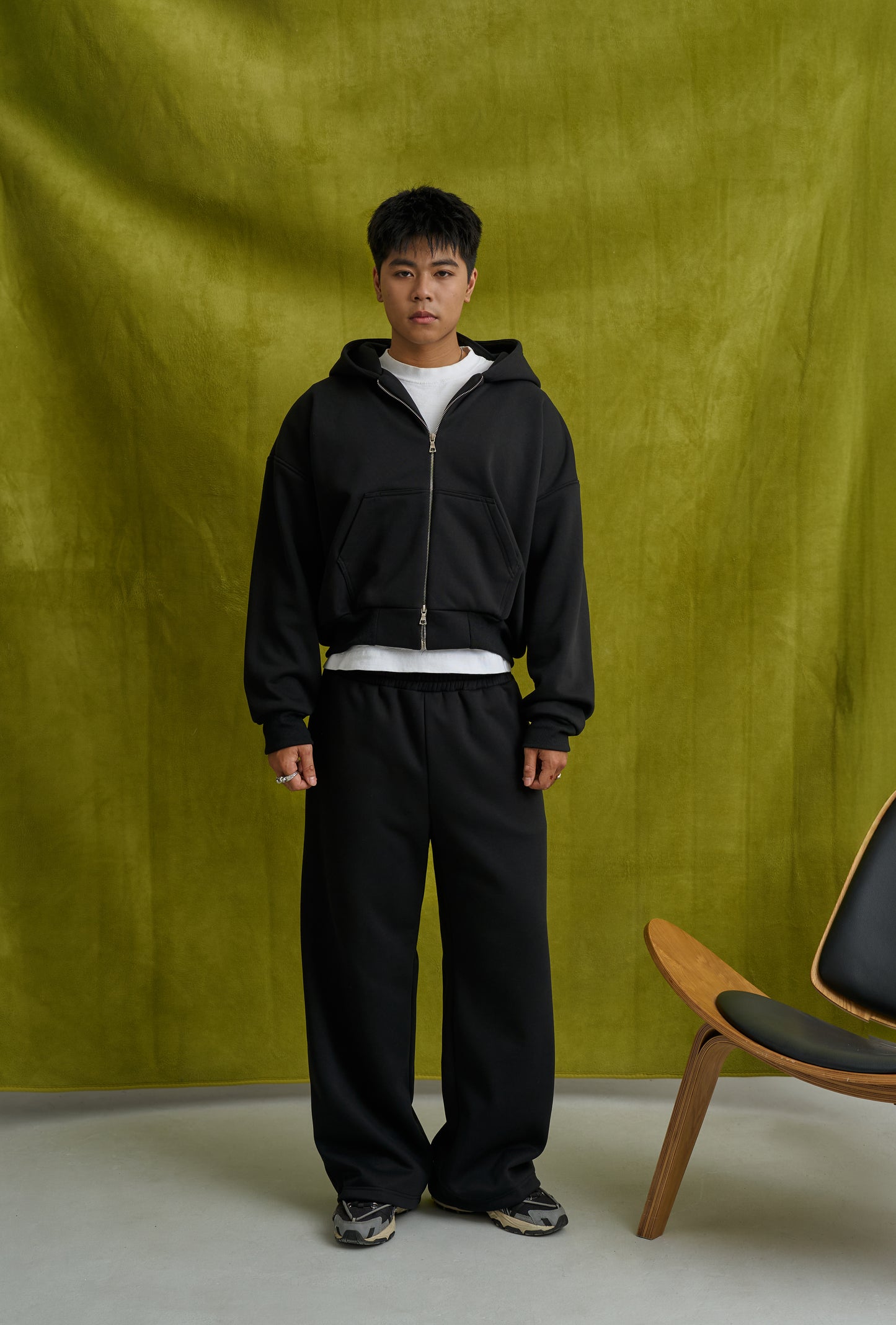 Person wearing a black tracksuit standing against a green backdrop with a chair visible.