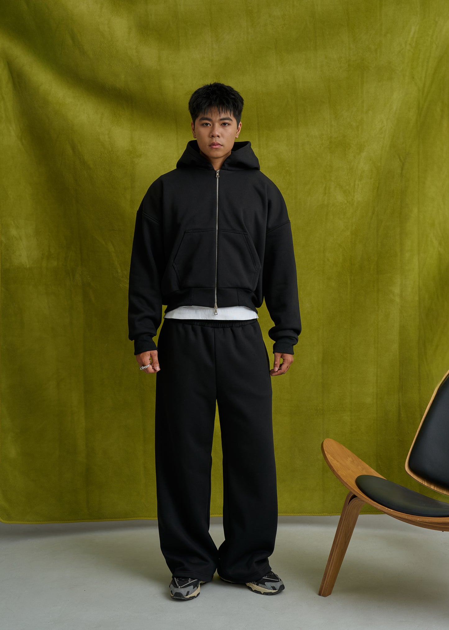 Person wearing a black hoodie and pants standing against a green curtain with a chair in the corner.