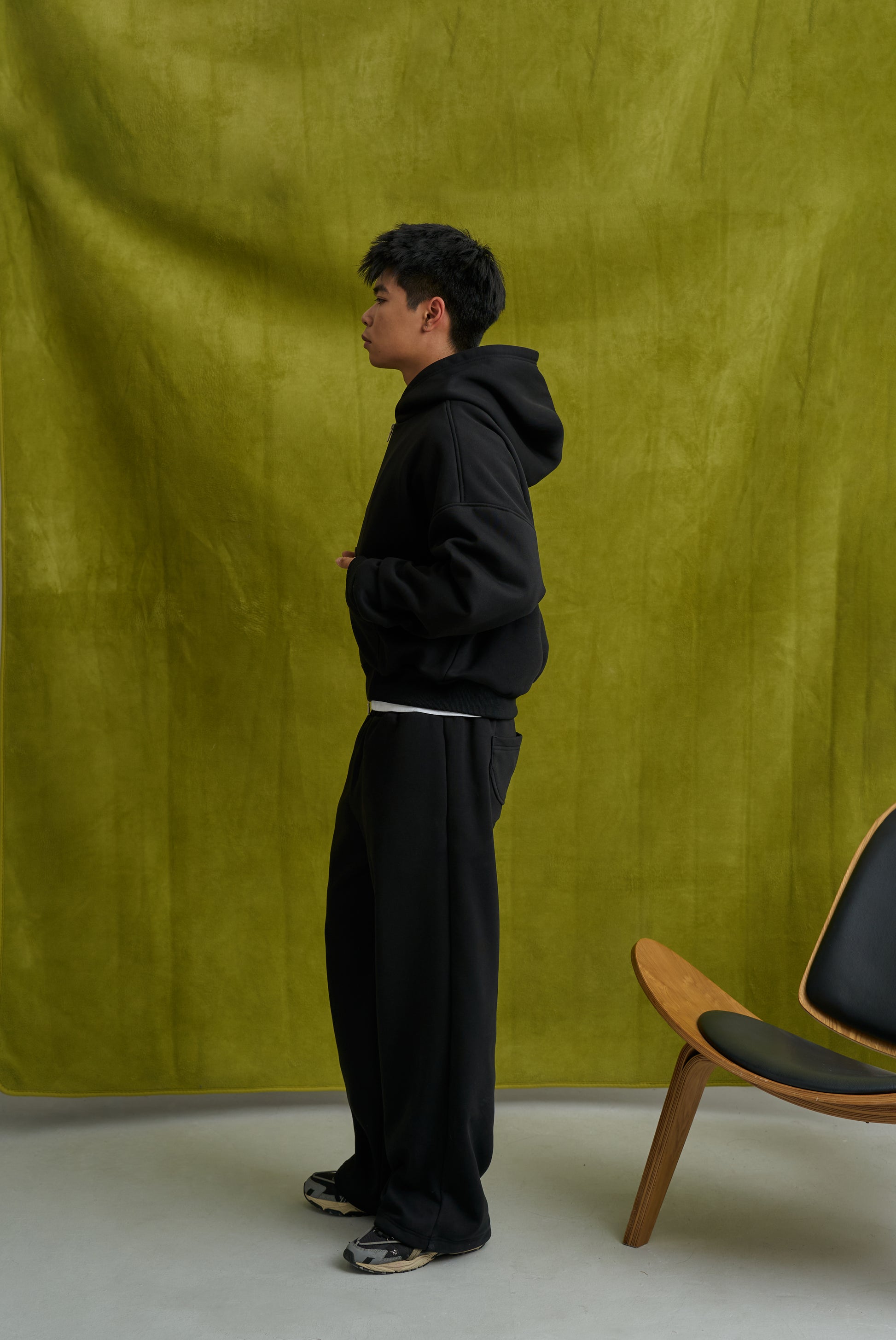 Person wearing a black hoodie and pants standing against a green curtain with a chair in the foreground.