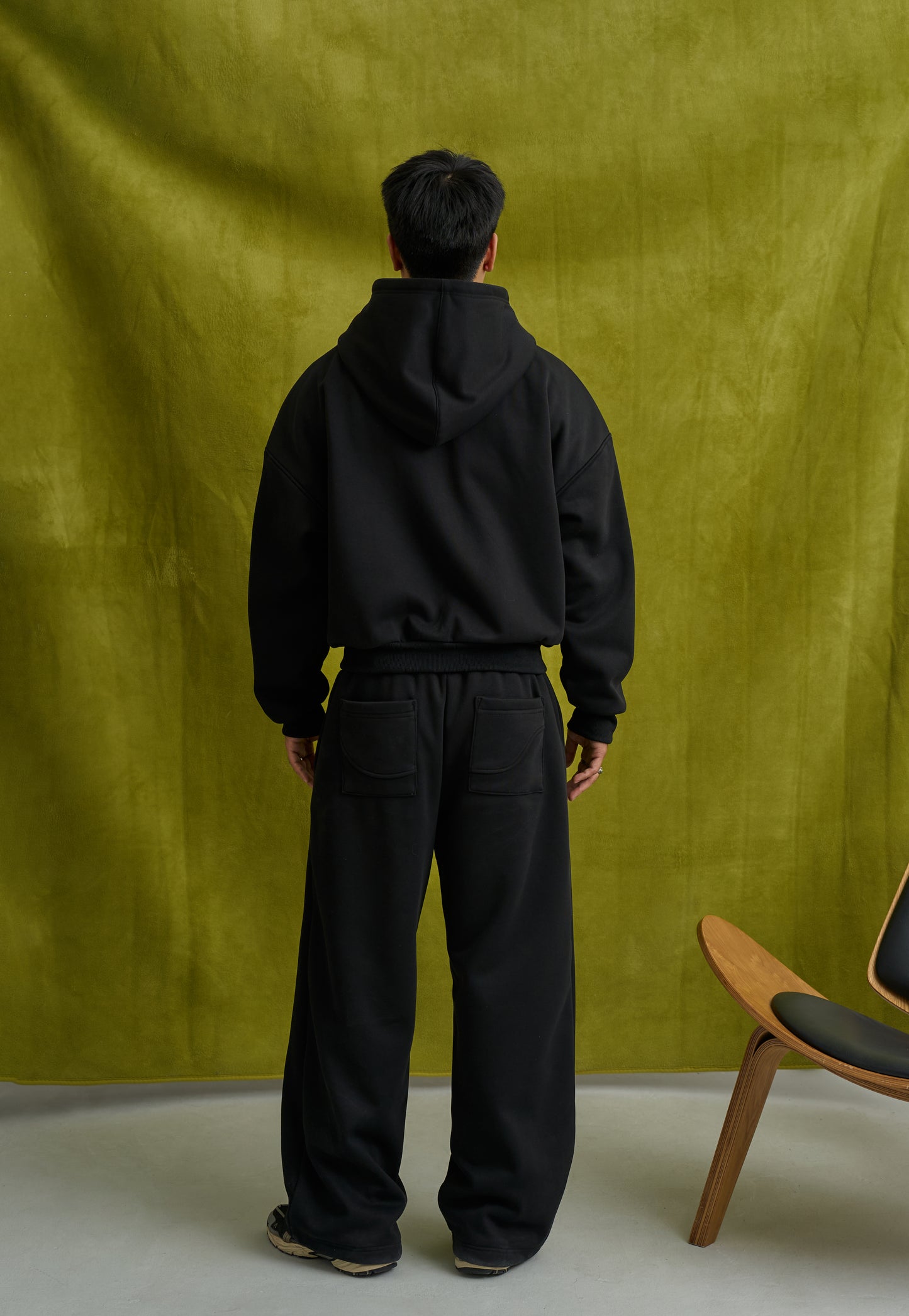 Person wearing a black hoodie and pants standing against a green curtain with a chair on the side.