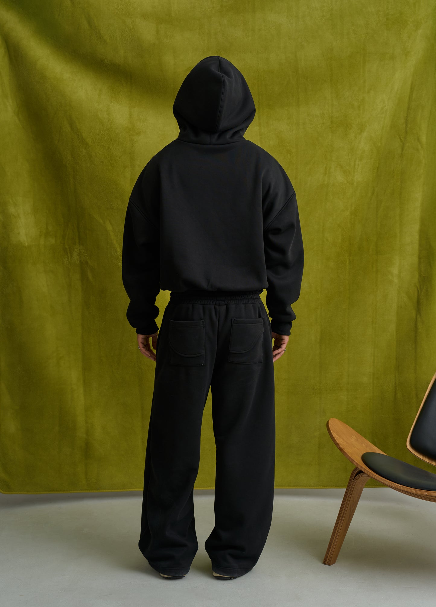 Person wearing a black hoodie and pants standing against a green curtain with a chair in the corner.